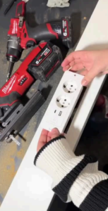 Close-up of hands holding a white inbuilt power outlet with USB ports, next to red Milwaukee power tools during a DIY bedroom installation.