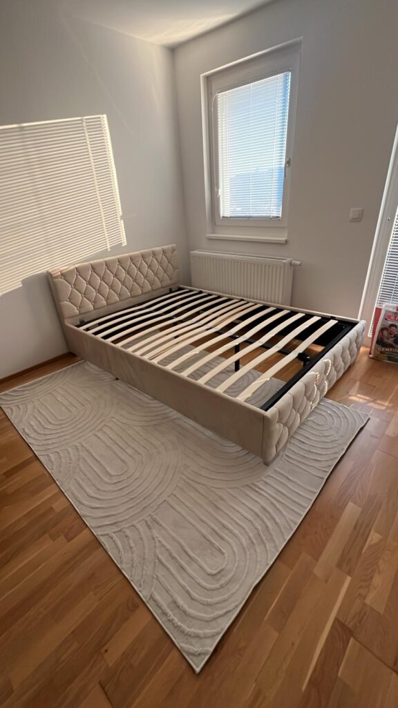 SIMPEX beige boho rug with 3D geometric design placed under a bed frame in a modern bedroom with wooden flooring.